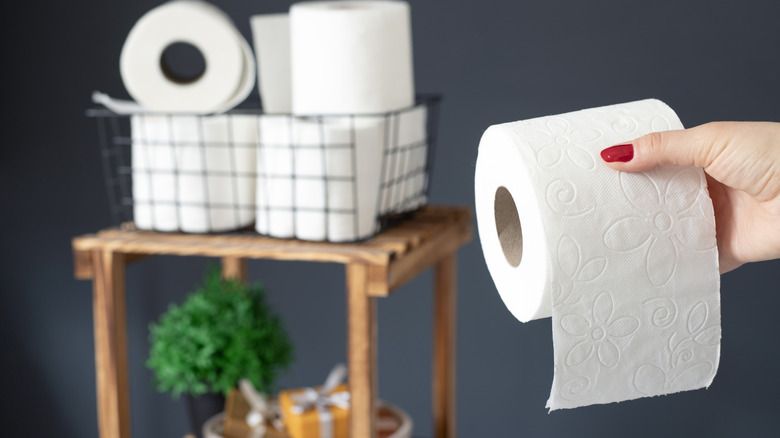 A hand holds up a roll of toilet paper, with a basket containing other rolls in the background