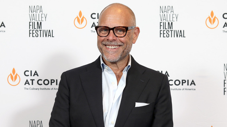 Alton Brown smiling in front of a promotional background at an event