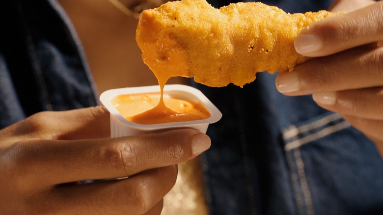 A woman holds a McCrispy Strip in one hand as she dunks it into a spicy orange dipping sauce with the other
