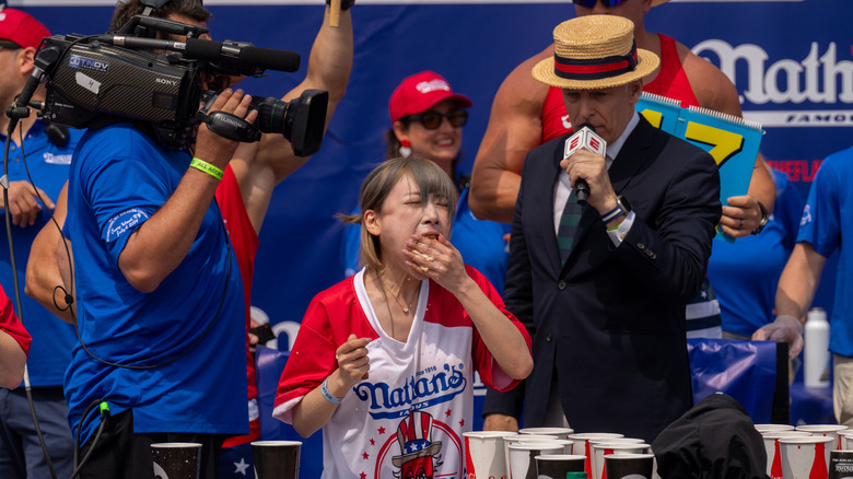 A competitive eater shoves a hot dog in her mouth while an announcer and cameraman stand over her