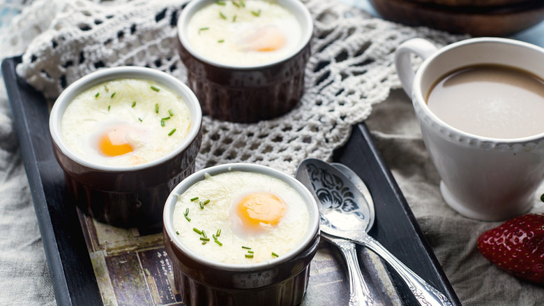 Three shirred eggs in black ramekins on a black tray with a lace doily and two teaspoons with a cup of coffee and a strawberry on the side