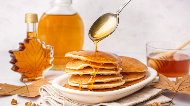 Maple syrup bottles sit in the background as a spoon drips syrup onto a stack of pancakes in the foreground