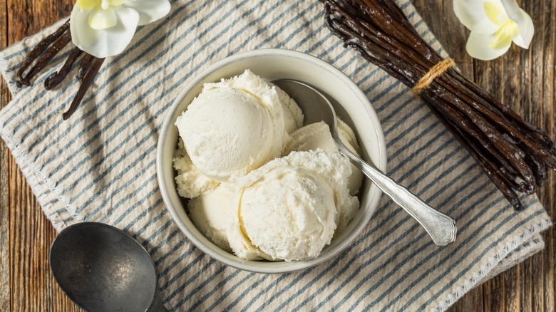 A bowl of vanilla ice cream sitting on a kitchen towel with a spoon and whole vanilla beans on the side