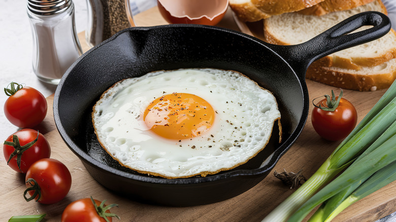 A fried egg in a skillet on a table next to cherry tomatoes, sliced bread, scallions and salt and pepper