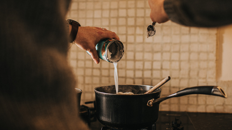 Hand pours coconut milk from a can into a pot on the stove