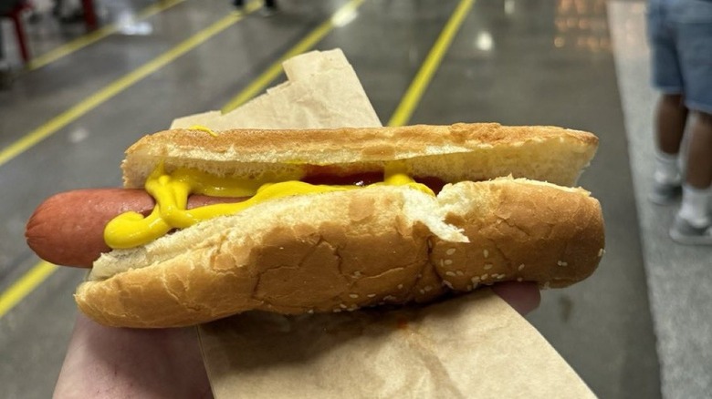 A man holds out a Costco hot dog topped with mustard and ketchup