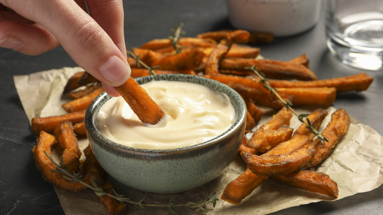 A hand dipping sweet potato fries into a pot of mayo.