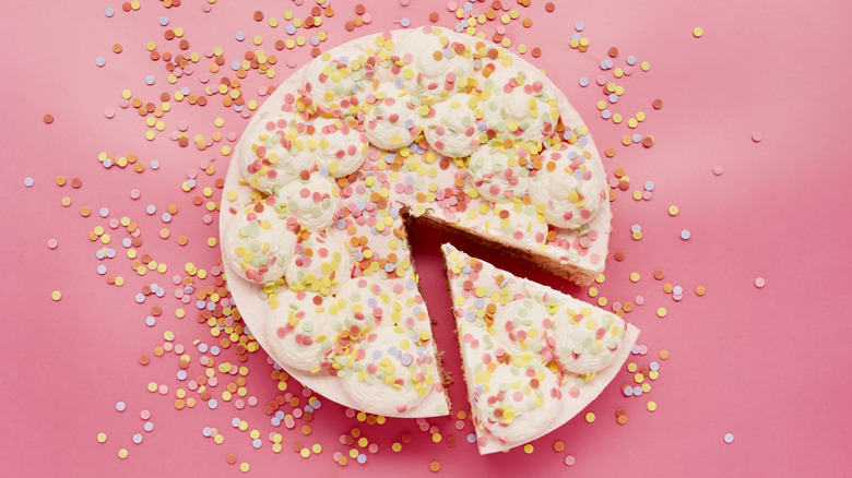 A cake with confetti sprinkles has a slice slightly cut out, against a pink background