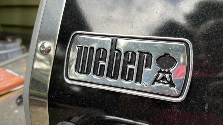 A close-up of a Weber gas grill's logo on the front of the grill hood.