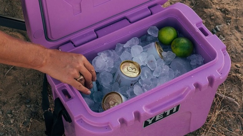 A hand reaches into a Yeti cooler packed with ice, cans, and limes