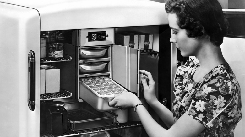 In black and white, a homeowner puts a tray of ice cubes into a vintage refrigerator