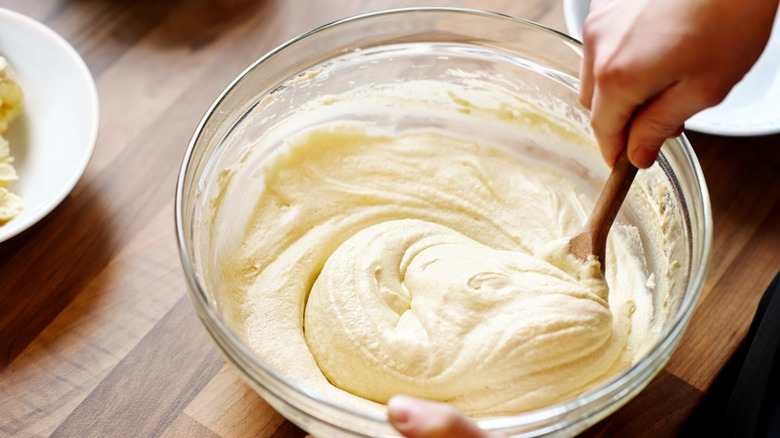 A woman uses a wooden spoon to mix a creamy batter in a glass bowl