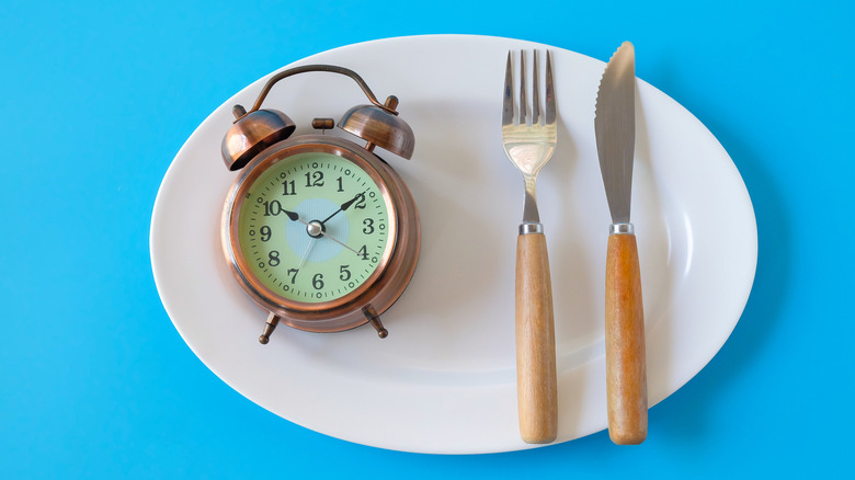 An alarm clock and a knife and fork set sit on an oval dinner plate