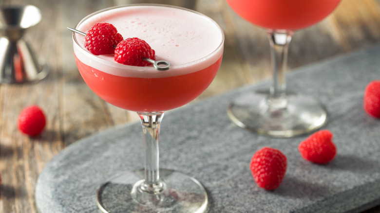 Reddish-pink cocktail in a martini glass topped with egg white foam and two raspberries for garnish