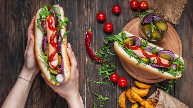 Someone holds a hot dog laden with toppings, next to a wooden plate containing another hot dog and a ramekin with gherkins and red onions, along with fried potato wedges, cherry tomatoes, herbs, and a chili pepper scattered across a wooden surface
