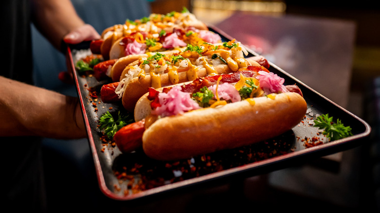 Person holds a tray of hot dogs in buns heaped with various toppings
