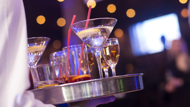 Server carrying silver tray of cocktails in various glasses.