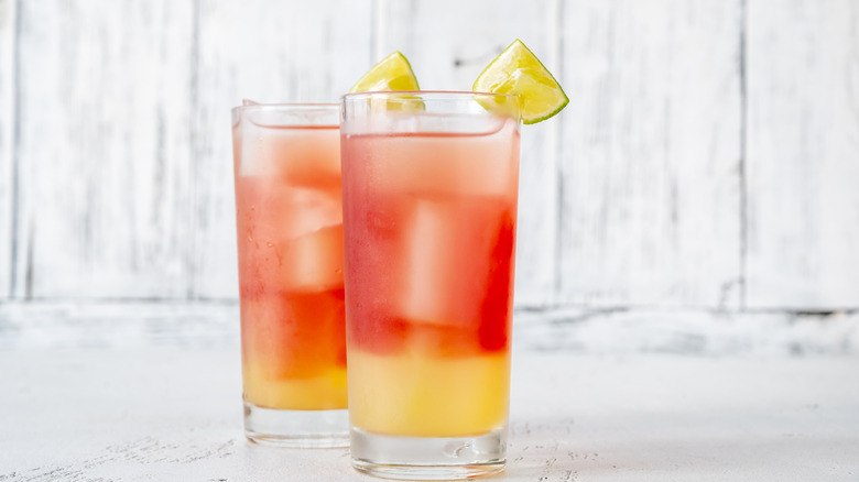 Two glasses of layered bay breeze cocktails, garnished with lime wedges.