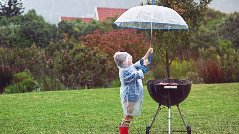 A smiling child holds an umbrella over a charcoal grill