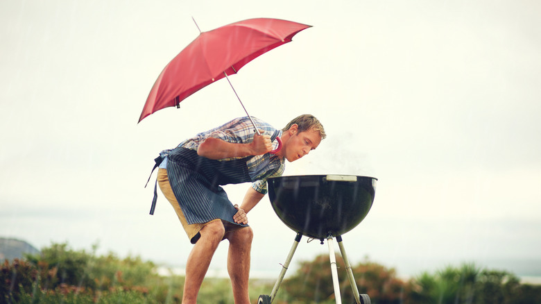 A man holding an umbrella bends over to blow on a charcoal grill in the rain