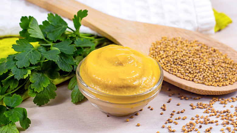 Dijon mustard in a small glass bowl next to parsley sprigs and a large wooden spoon holding mustard seeds