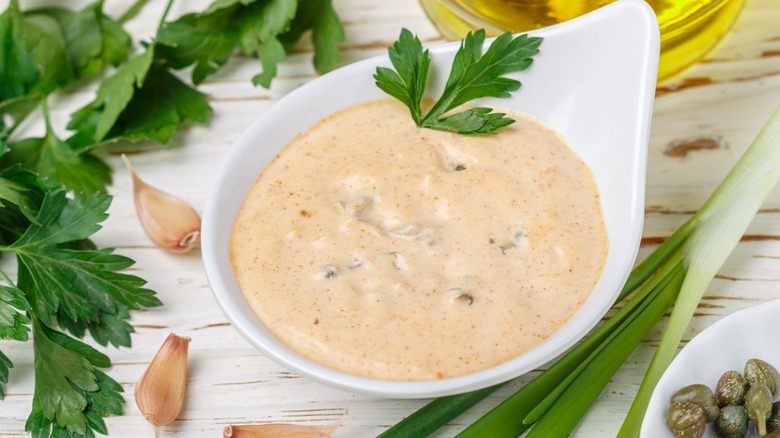 A French remoulade sauce in a white bowl with a sprig of parsley, surrounded by capers, garlic, and scallions.