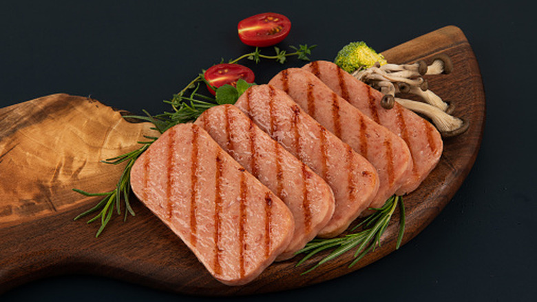 Grilled Spam on a board with veggies and fresh herbs alongside it.