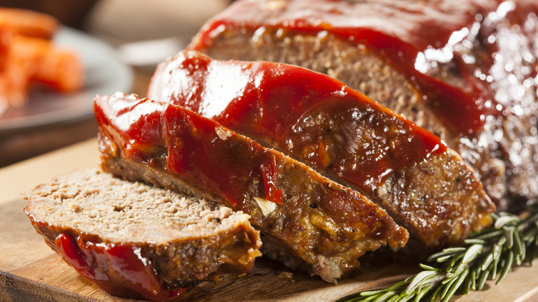 A sliced meatloaf on a cutting board with a sprig of fresh rosemary and a ketchup glaze