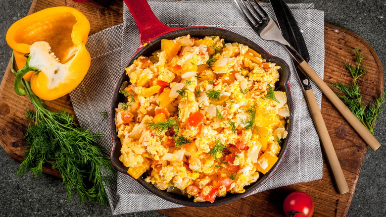 Scrambled eggs with red and yellow pepper pieces in a red-handled pan on wooden cutting board with a grayish kitchen towel on top, with a half-cut yellow pepper, fresh dill, a cherry tomato and a fork and knife on the side