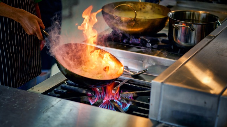 A cook in a kitchen holds a wok over high flames on a stove as fire leaps up