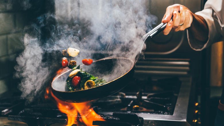 A chef tossing vegetables in a sloped pan over an open flame
