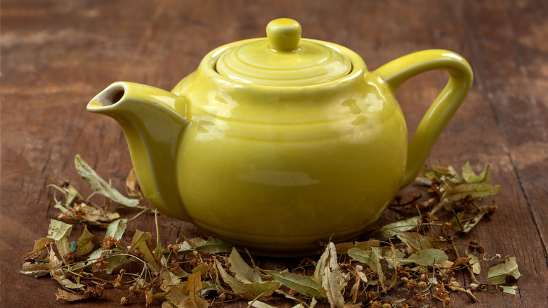 A cheerful yellow teapot sits on a table, surrounded by dried herbs