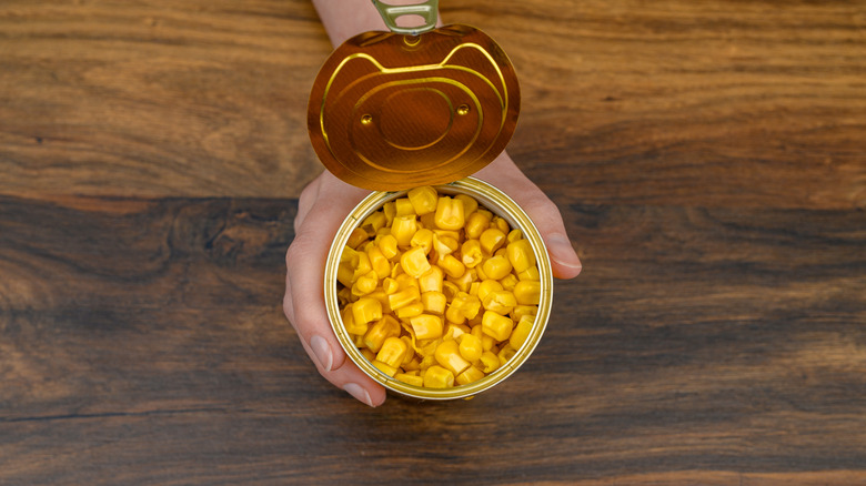 A hand holding an open can of corn on the table