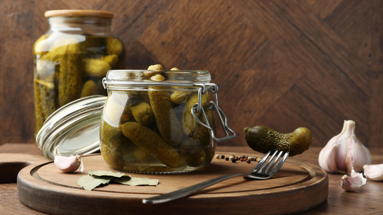 An open jar of pickled gherkins on a lazy Susan with garlic, bay leaves, and peppercorns and a sealed pickle jar in the background