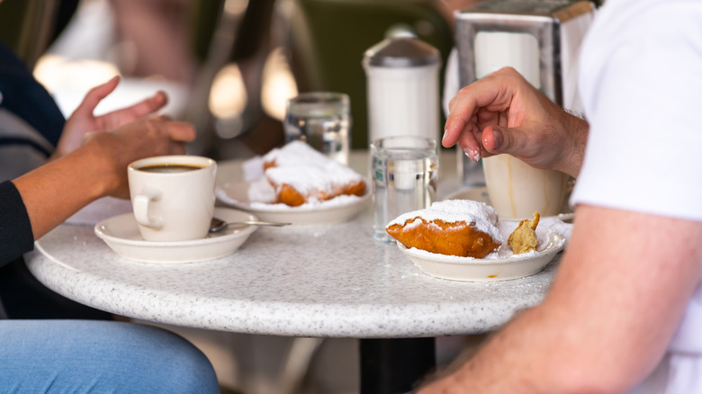 A couple sits at an outdoor table with coffee and beignets