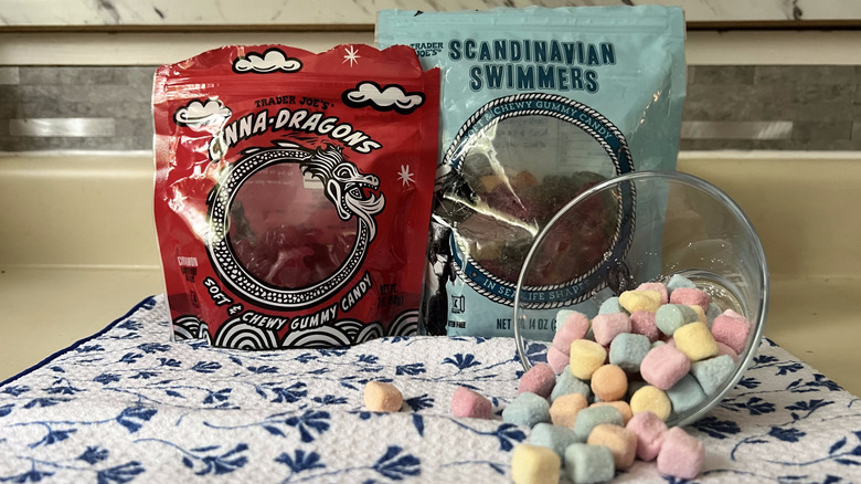 A bag of Cinna-Dragons next to a bag of Scandinavian Swimmers both of which are background to a turned over glass cup full of confetti chews