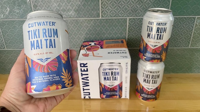 Hand holding a can of Cutwater's Tiki Rum Mai Tai with a case and two more cans in the background