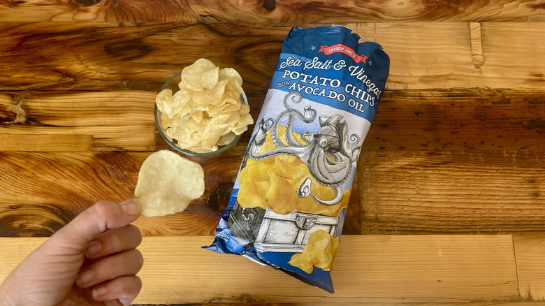 Overhead view of a bag and bowl of Sea Salt & Vinegar Potato Chips With Avocado Oil on a wooden table with a hand holding a single chip