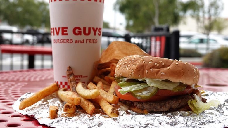 A burger and fries on an aluminum wrapper next to a drink on a table at a Five Guys location.