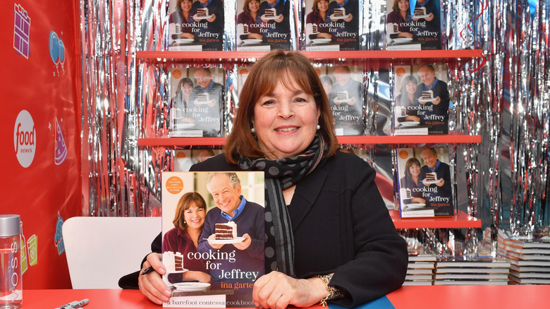 Ina Garten sitting at table for book signing, holding a cookbook with a wall of books behind her