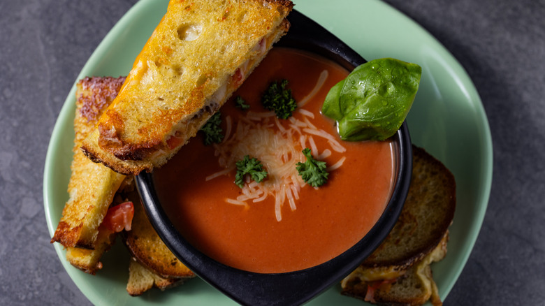 A grilled cheese sandwich sits next to a bowl of tomato soup garnished with fresh basil and parsley7#][