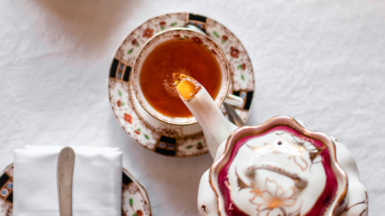 Someone pours black tea into a tea cup and saucer from a tea pot23.0