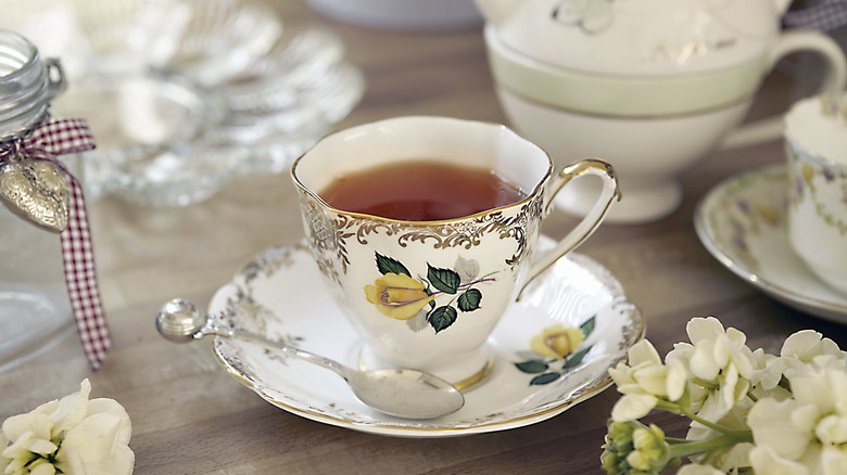 An English tea cup and setting featuring flowers is full of tea and sits beside fresh flowers