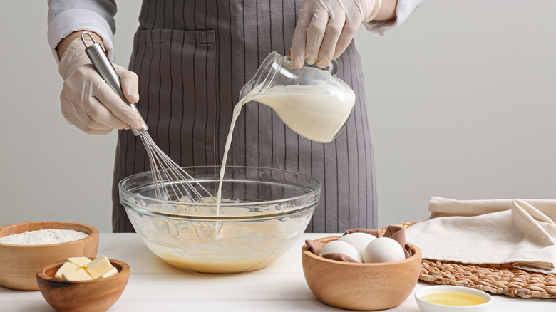 A man holds a whisk in one hand while pouring milk into a bowl for batter