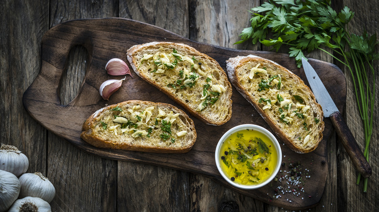 Top view of three slices of garlic bread with garlic butter, parsley, garlic cloves, salt, and seasonings all on a wooden table and cutting board.