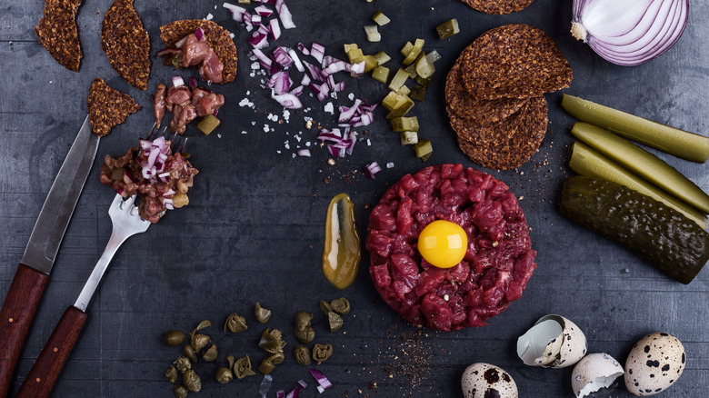 A round of steak tartare sits in the midst of accoutrements, such as pickles, onions, and quail eggs