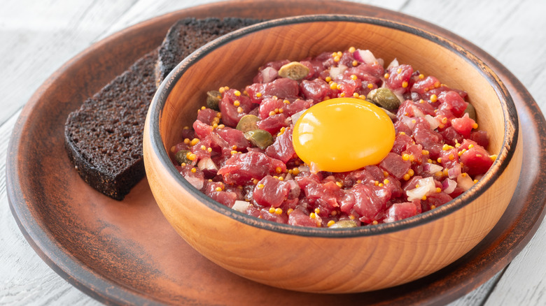 Steak tartare, topped with an egg yolk, sits in a bowl on a plate with bread