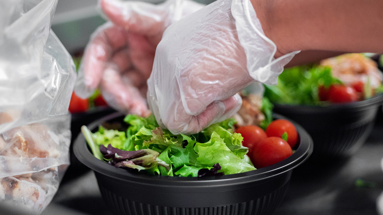 Two gloved hands place fresh salad ingredients in bowls