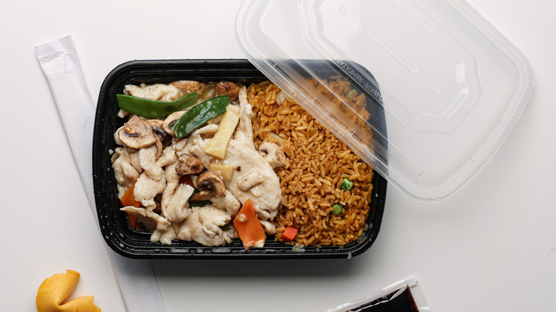 A Chinese takeout meal with stir-fried chicken, vegetables, and fried rice in a black plastic container, with a fortune cookie, soy sauce packet, and disposable chopsticks on a white background