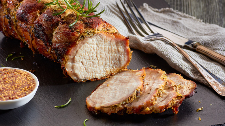 Sliced cooked pork loin with rosemary on a black surface next to bowl of whole mustard seeds
